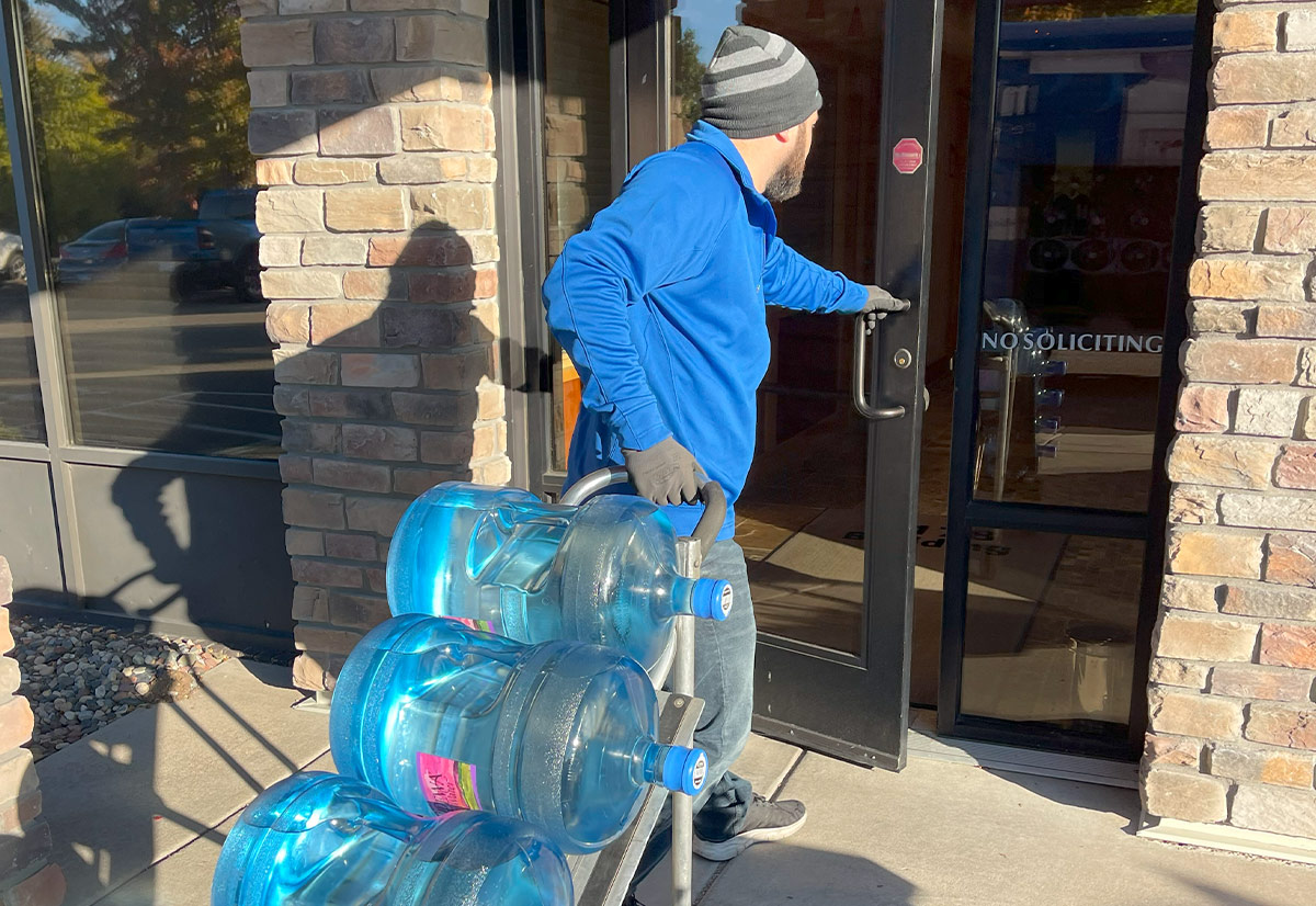A delivery driver in a blue jacket and knit hat pulling a hand truck stacked with three five gallon water bottles through the front door of a brick office building