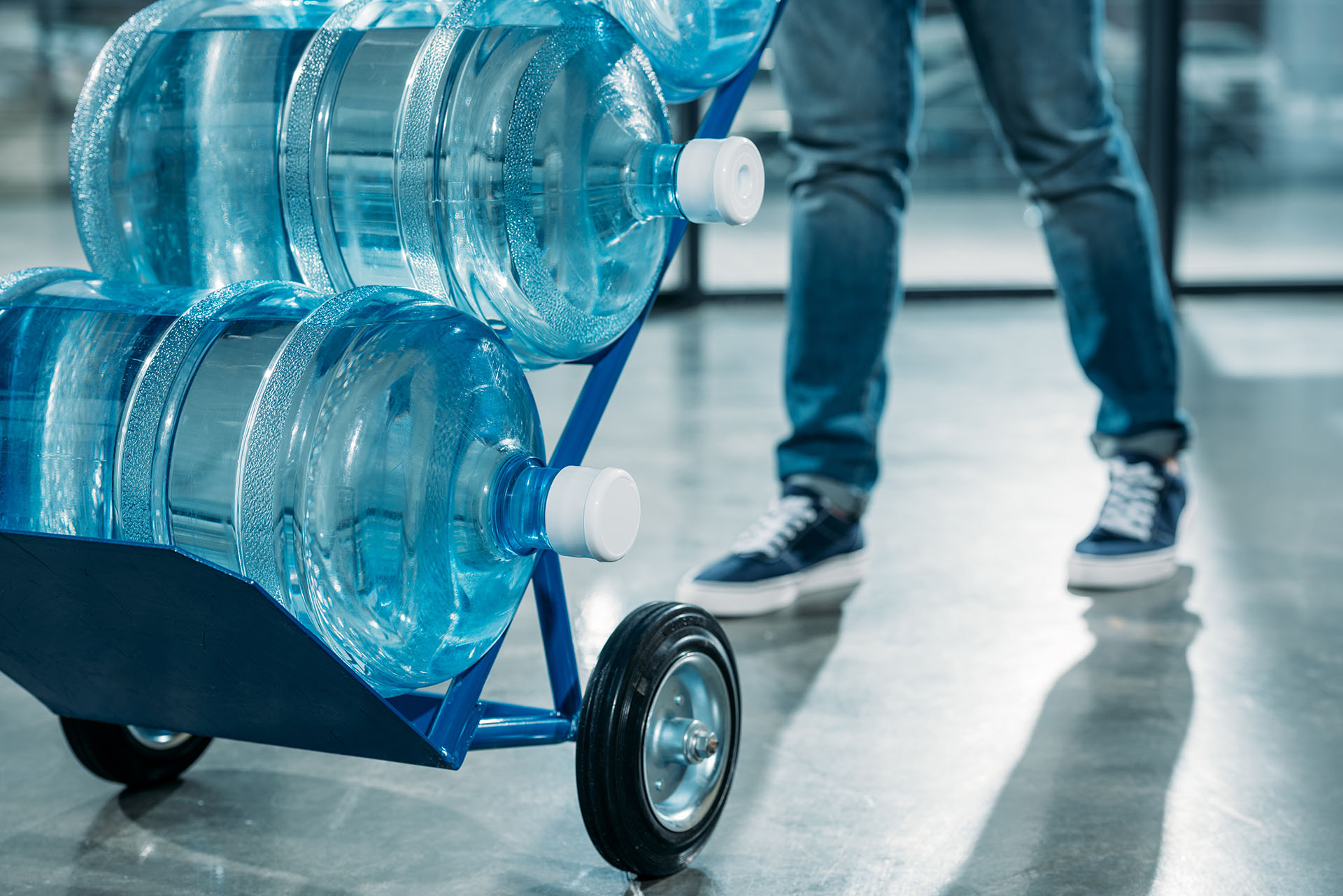 A blue hand truck loaded with four five gallon water bottles being wheeled across a polished floor, with the delivery person walking behind it